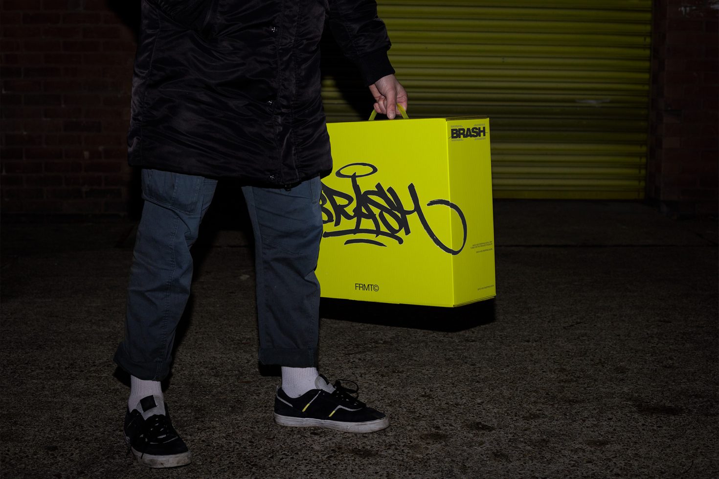 Person in urban setting holding vibrant yellow box with bold black graffiti-style design, showcasing packaging mockup for product branding.